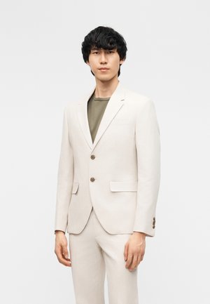 Młody mężczyzna o ciemnych, kręconych włosach, ubrany w kremową marynarkę i pasujące spodnie, nałożone na oliwkowozieloną koszulę, stojący na jednolitym tle.