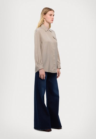 Béžová košile na knoflíky s límcem, dlouhými rukávy a přední kapsou, v kombinaci s volnými tmavě modrými džíny. Hladká textura, casual styl.