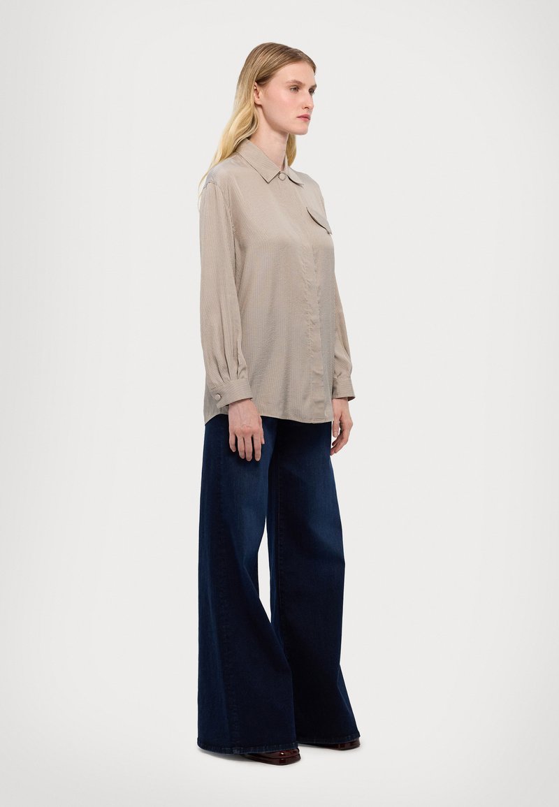 Beige button-up shirt with a collar, long sleeves, and a front pocket, paired with wide-leg dark blue jeans. Smooth texture, casual style.