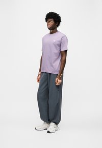 Ανοιχτό μωβ κοντομάνικο μπλουζάκι με μικρό λογότυπο, συνδυασμένο με φαρδιά σκούρα γκρι παντελόνια, ολοκληρωμένο με λευκά αθλητικά παπούτσια.