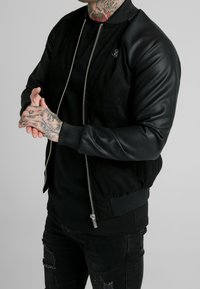 Svart bomberjacka med skinnärm och strukturerad kropp, med en silverdragkedja och ribbad mudd. Bärs med en svart skjorta under.