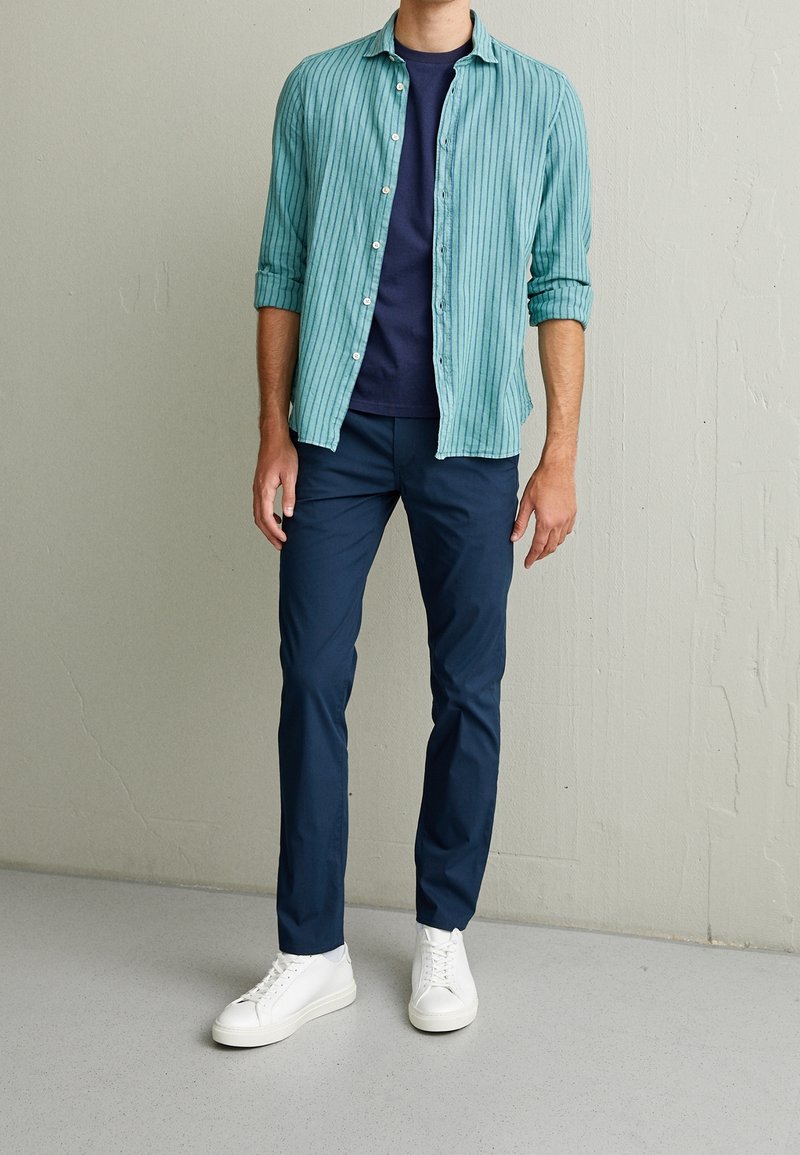 Homme portant une chemise rayée bleu sarcelle sur un t-shirt bleu marine, pantalon bleu marine et baskets blanches, debout contre un mur uni.