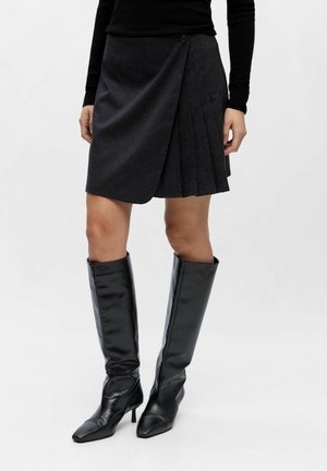 Jupe plissée gris foncé avec un design portefeuille, dotée d'une fermeture à boutons et d'un tissu texturé. Associée à des bottes noires brillantes montant jusqu'aux genoux.