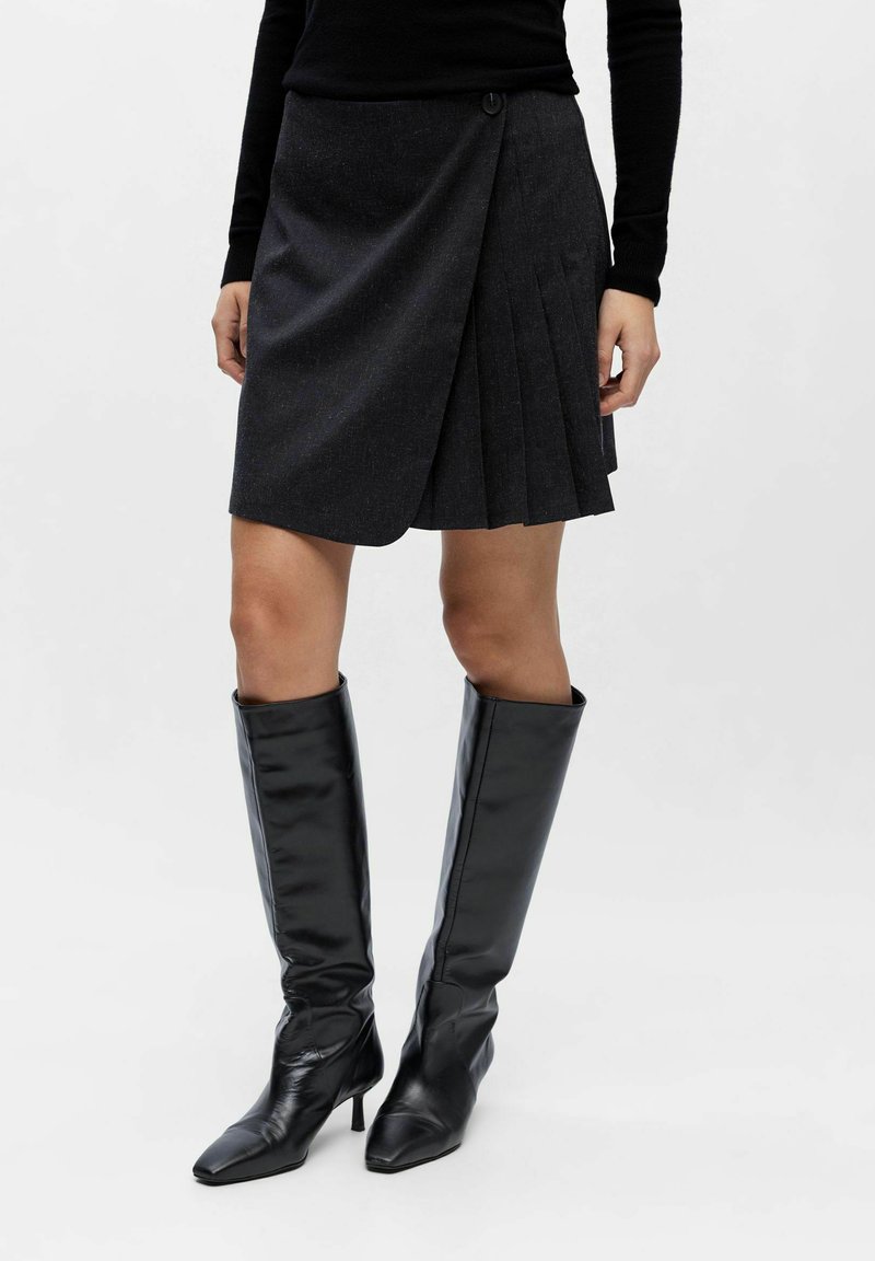 Mørkegrå plisseret nederdel med omslagsdesign, der har lukning med knapper og en struktureret stof. Parret med skinnende sorte knæhøje støvler.