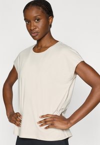 Ανοιχτό μπεζ αθλητικό τοπ με κοντά μανίκια και στρογγυλή λαιμόκοψη. Το ύφασμα φαίνεται λείο και ελαστικό, με χαλαρή εφαρμογή και διακριτική λεπτομέρεια λογότυπου.