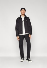 Sort zip-up jakke med høj krave og logo på brystet, båret over en hvid t-shirt, parret med sorte bukser og sneakers.