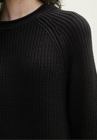 Pull en maille noire avec texture, présentant un col rond et des détails côtelés aux manches, offrant une coupe décontractée.