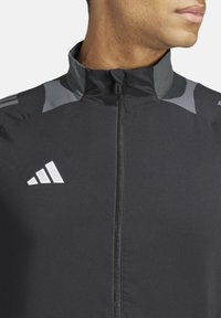 Giacca sportiva nera con colletto alto, con dettagli grigi e un logo bianco. Realizzata in tessuto liscio e leggero, con una zip frontale.