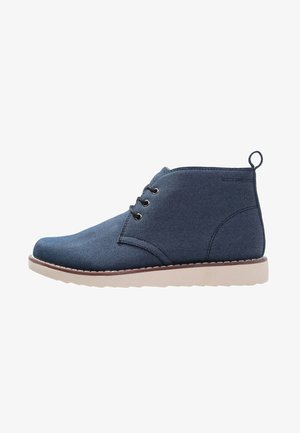 Bottes cheville en denim bleu avec une semelle en caoutchouc crème, deux œillets et une boucle au talon. Présente une texture lisse et un design à bout arrondi.
