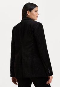 Blazer in corduroy nero con vestibilità su misura, revers a ribalta e polsini a tre bottoni. Texture liscia, spaccone singolo sul retro e design minimalista.