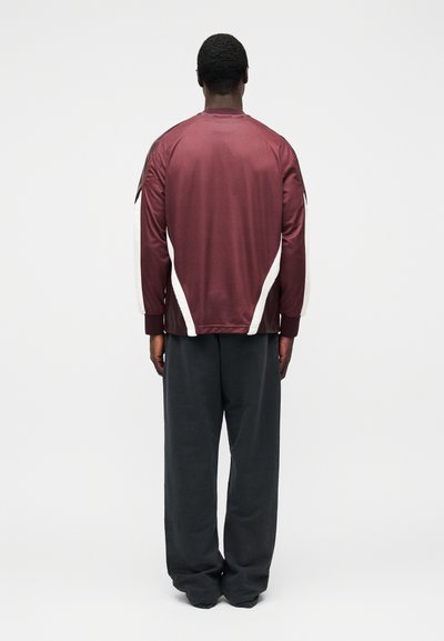 Bordo värvi sportlik jakk, millel on valged küljekaunistused, seisev krae ja pikad varrukad. Lahtised tumedad hallid püksid, millel on sile tekstuur.