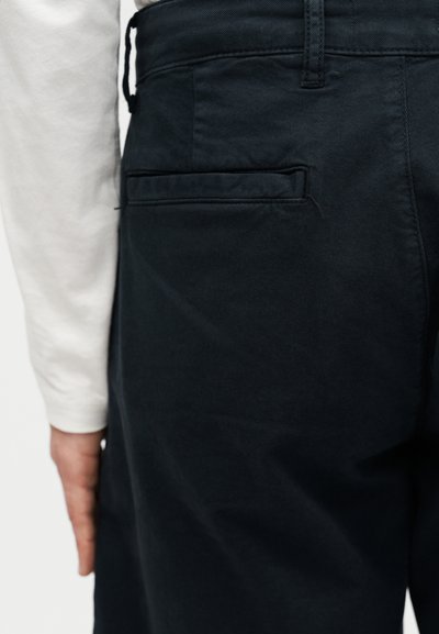 Κοντινό πλάνο ενός ατόμου που φοράει μαύρο παντελόνι με πίσω πλαϊνή τσέπη και λευκό πουκάμισο με μακριά μανίκια, με το χέρι ορατό στο πλάι.