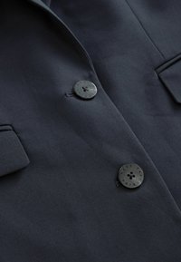 Marine blazer med tekstureret stof, to store sorte knapper og notch-lapel. Enkel design med minimale hardware-detaljer.
