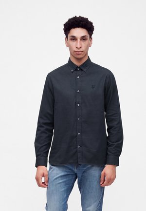 Junger Mann mit kurzen lockigen Haaren, der ein schwarz geknöpftes langärmeliges Hemd und hellblaue Jeans trägt, steht vor einem schlichten weißen Hintergrund.