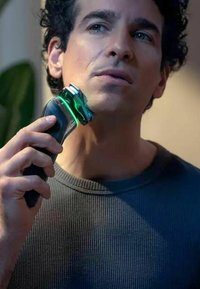 Rasoio elettrico con corpo nero e dettagli verdi, dotato di una testina a più lame. Utente che lo tiene contro la linea della mascella, indossando una maglietta grigia.