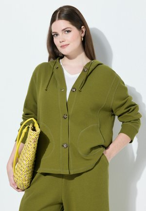 Olijfgroene gebreide hoodie met een voorste knoopsluiting, grote zakken en contrasterende stiksels. Geweven gele handtas met een gestructureerd patroon.