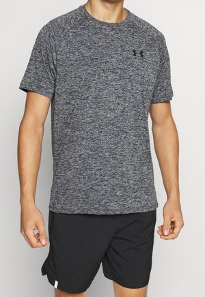 Mężczyzna ubrany w szary sportowy t-shirt z krótkim rękawem i czarne spodenki stoi na jednolitym tle.