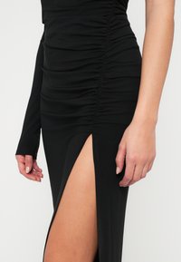 Crna haljina s nabranim materijalom, jednim dugim rukavom i visokim prorezom do buta. Glatka tekstura sa uskim siluetom i čistim detaljima šavova.