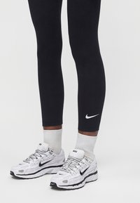 Άτομο που φοράει μαύρα κολάν Nike, λευκές κάλτσες Nike και λευκά παπούτσια για τρέξιμο Nike με μαύρες λεπτομέρειες, στέκεται σε ανοιχτό φόντο.