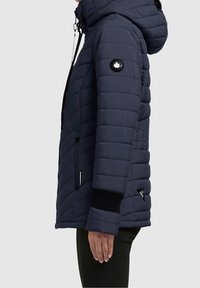 Granatowa kurtka puchowa z kapturem, pikowane wzornictwo, zamykane kieszenie i ściągacze na rękawach. Posiada logo naszywkę na lewym rękawie.