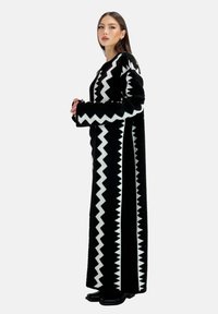 Long manteau noir avec un motif en zigzag blanc, confectionné dans un tissu texturé, doté de larges manches et d'une fente latérale, associé à des chaussures noires.
