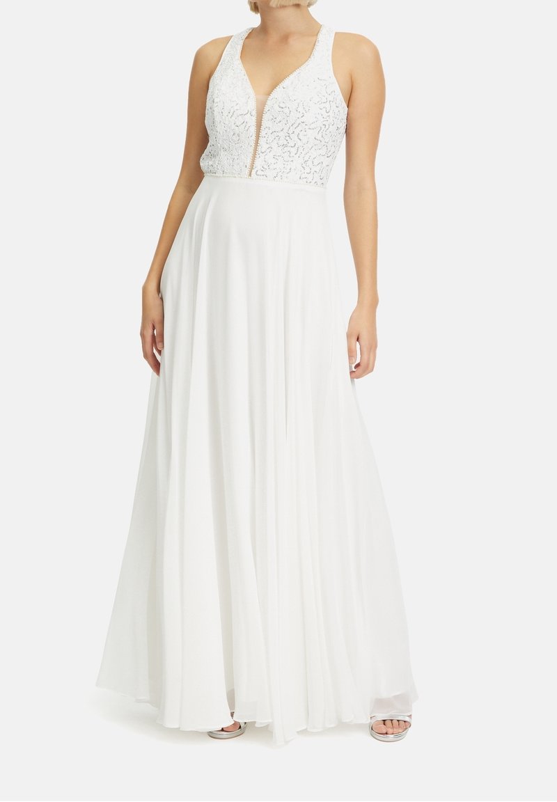 Femme portant une robe blanche sans manches avec un corsage en dentelle à décolleté en V profond et une longue jupe fluide, debout sur un fond clair uni.