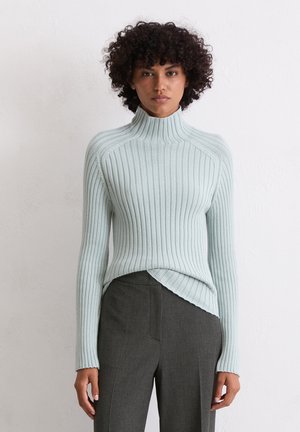 Vrouw met krullend haar, gekleed in een lichtblauwe geribbelde coltrui en grijze geblokte hoog-waisted broek tegen een effen muur.