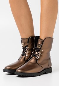 Botines metálicos marrones con cordones negros, forro de pelaje sintético y acentos de hardware decorativo en los lados. Suelas planas negras.