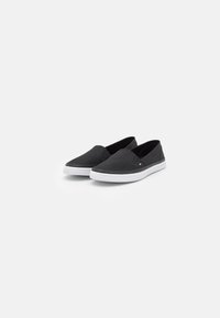 Ein Paar schwarze Leinenschuhe mit Gummisohlen in Weiß, ausgestattet mit einem kleinen Logo-Flag auf der Seite, vor einem weißen Hintergrund.