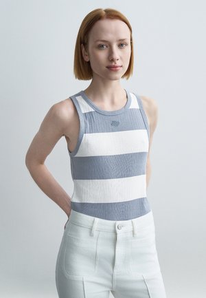 Débardeur à côtes avec des rayures horizontales bleues et blanches, doté d'un col rond et d'un petit logo sur la poitrine. Un pantalon blanc complète la tenue.