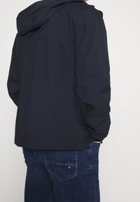 Mörkblå huva jacka med en slät, matt yta, som har en enkel design och elastiska manschetter. Bärs med mörka denimjeans.