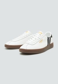 Bílé sneakers vyrobené z kůže, s hnědou gumovou podešví, černými proužky po stranách a zlatým logem na jazyku.