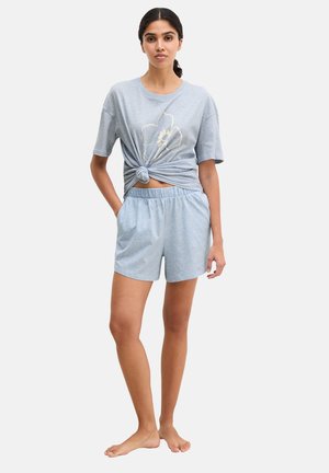 Kobieta stojąca boso, ubrana w jasnoniebieskie szorty i krótki, wiązany na supeł t-shirt z dużym kwiatowym wzorem.