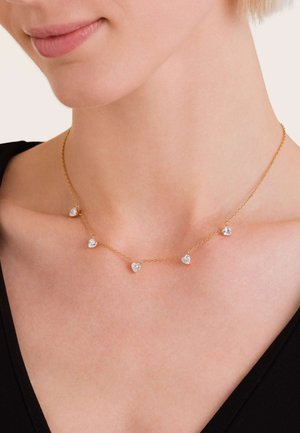 Gouden ketting met vijf hartvormige accenten met heldere stenen, tentoongesteld op een halslijn tegen een zwarte kledingstuk.