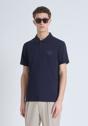 Navyblauw poloshirt gemaakt van zachte stof, met een drieknopenlijst en een reliëflogo op de linkerkant van de borst. Gekleed met lichtgekleurde broeken.