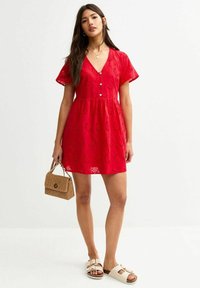 Rotes Baumwollkleid mit kurzen, fließenden Ärmeln, V-Ausschnitt, Knöpfen, gestickten Details und einem ausgestellten Rock. Getragen mit einer Strohtasche und Sandalen.