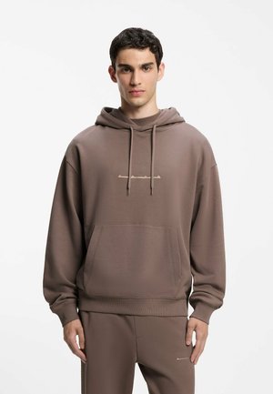 Jonge man die een bruine hoodie en bijpassende broek draagt met een voorzak en trekkoorden, staand tegen een effen witte achtergrond.