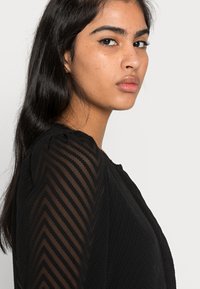 Top preto com tecido texturizado e padrões em zigzag, decote redondo e mangas longas transparentes. Design liso e ajustado com detalhes subtis.