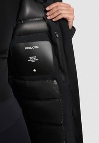 Fekete puffernadrág, sima külső felülettel, cipzáras zsebbel és logóval ellátott folttal. A belső rész steppelt szigetelést mutat a melegség érdekében.