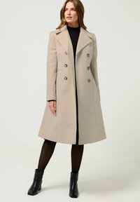 Beige wollen jas met een dubbelknopsdesign, grote revers, zilveren knopen en een gestructureerde vorm. Draag het over een zwarte coltrui.