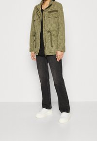 Olijfgroene geblokte jas met twee borstzakken en een trekkoord in de taille, gecombineerd met zwarte jeans en witte sneakers.