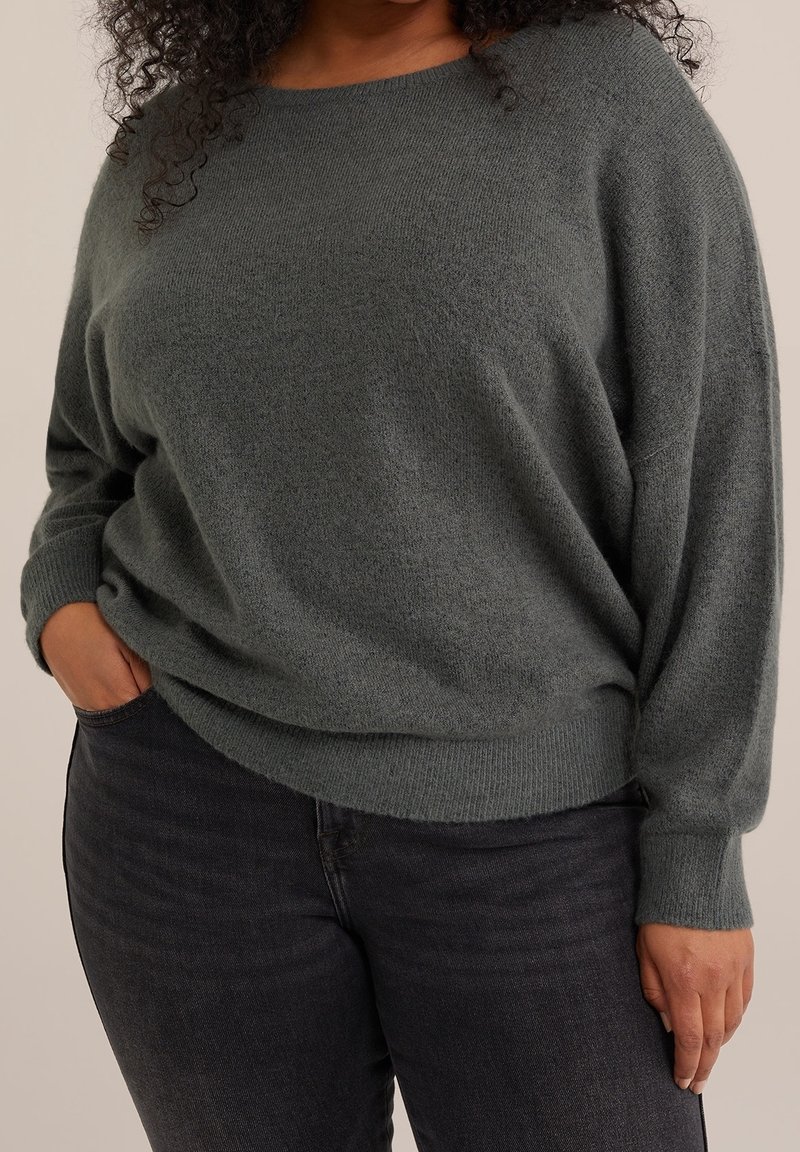 Femme portant un pull gris ample à manches longues et un jean foncé, une main dans la poche sur un fond neutre.