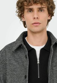 Grau-profiliertes Fischgrätmuster-Jackett mit Kragen und Knopfdetails, über einem schwarzen gerippten Reißverschluss-Pullover getragen, der über einem weißen Hemd getragen wird.