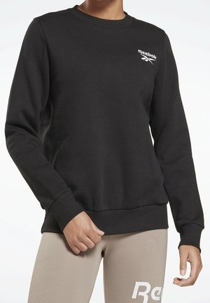 Osoba ubrana w czarną bluzę Reebok z logo na piersi oraz beżowe spodnie z białym napisem Reebok na udzie, stojąca na jednolitym tle.
