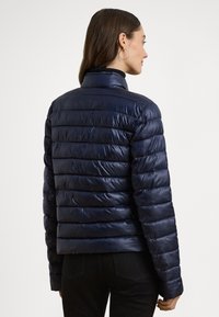 Μπλε σκούρο μπουφάν τύπου puffer με ραμμένη σχεδίαση και οριζόντιες ραφές, ψηλό γιακά και στενή εφαρμογή, κατασκευασμένο από γυαλιστερό, ελαφρύ υλικό.