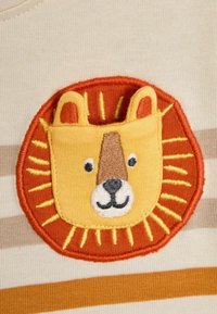 Kreemivärvi kangas pruunide ja oranžide triipudega, millel on tikitud lõvi näo patch, mille ümber on päikesepaiste muster.