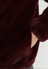 Burgundowy materiał przypominający futro, miękka tekstura, długie rękawy, ręka w bocznej kieszeni, charakteryzujący się puszystym, grubym designem.