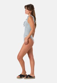 Femme debout de profil portant un maillot de bain une pièce rayé à volants et des sandales à plateforme noires sur un fond uni.