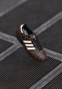 Sneaker a serpente marrone con lacci neri, strisce bianche e suola nera. Logo visibile sulla linguetta. Parte superiore in tessuto e parte inferiore in gomma.