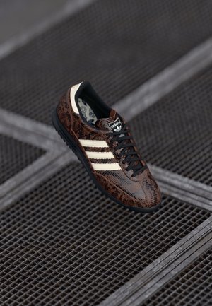 Sneaker a serpente marrone con lacci neri, strisce bianche e suola nera. Logo visibile sulla linguetta. Parte superiore in tessuto e parte inferiore in gomma.